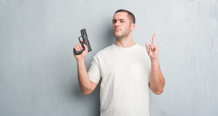 Young caucasian criminal man over grey grunge wall holding gun surprised with an idea or question pointing finger with happy face, number one