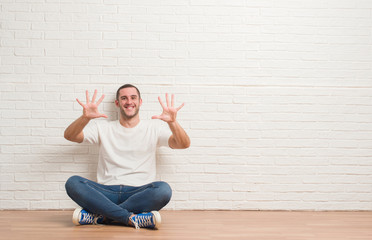 Young caucasian man sitting on the floor over white brick wall showing and pointing up with fingers number ten while smiling confident and happy.