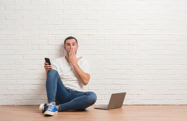 Young caucasian man sitting over white brick wall using computer laptop and smartphone cover mouth with hand shocked with shame for mistake, expression of fear, scared in silence, secret concept