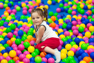 Fototapeta na wymiar Pretty small girl plays in children zone in amusement mall, lies on colourful balls, smiles positively, looks at camera. Little child with two pony tails plays with colored balls. Childhood concept