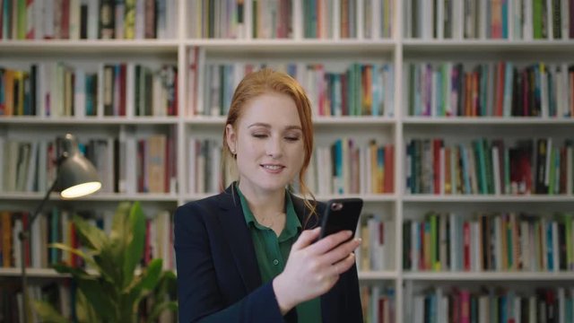 portrait of beautiful young red head business woman executive texting browsing using smartphone mobile app in library background