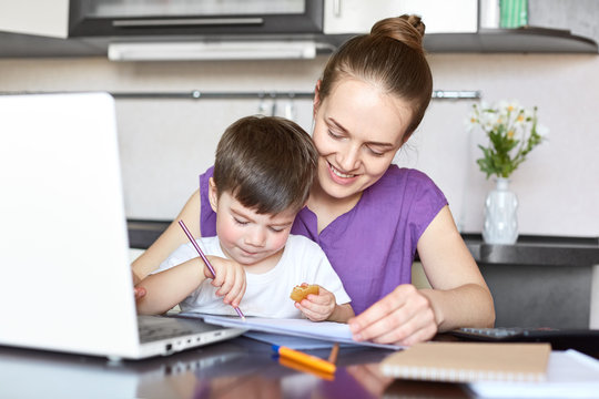 Cheerful mother cares of her son, being on maternity leave, works freelance with laptop computer, pose together at kitchen. Busy mom plays wit small boy, has rest after work. Family concept.