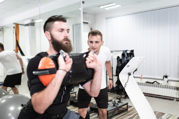 Fototapeta na wymiar Young bearded man doing exercises in electrical muscular stimulation suit with her personal trainer at rehabilitation center.