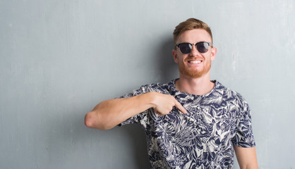 Young redhead man over grey grunge wall wearing sunglasses with surprise face pointing finger to...
