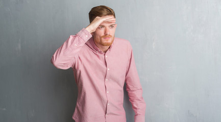 Young redhead man over grey grunge wall wearing pink shirt very happy and smiling looking far away with hand over head. Searching concept.