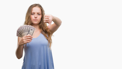 Young blonde woman holding dollars with angry face, negative sign showing dislike with thumbs down, rejection concept