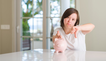 Down syndrome woman at home holding piggy bank with angry face, negative sign showing dislike with...