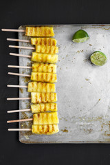 Grilled pineapple on bamboo sticks with lime on tray over black table, top view. From above, flat lay, overhead.