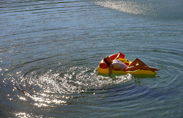 A young woman relaxes on an inflatable sun bed  in the middle of a lake. She wears a white swim ...