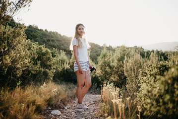 Cute girl posing on the sandy road  a lot of green plants with green hills and the sunset on the background in Spain. Traveler in the mountains.