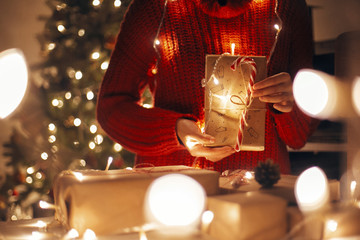 merry christmas. hands holding christmas present in lights in evening festive room under tree...