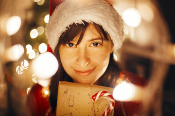 merry christmas. girl in santa hat and red sweater holding christmas present in lights in evening...