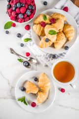 Sweet summer dessert, homemade baked mini croissants with berry jam, served with tea, fresh raspberries, blueberries and mint. On a white marble table, copy space top view