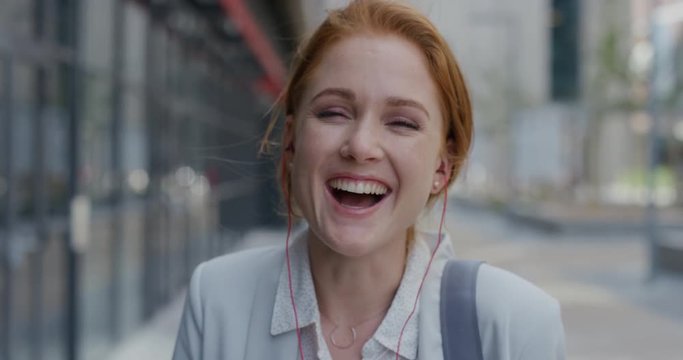 portrait happy young red head business woman laughing wearing earphones enjoying relaxed urban lifestyle listening to music in city slow motion