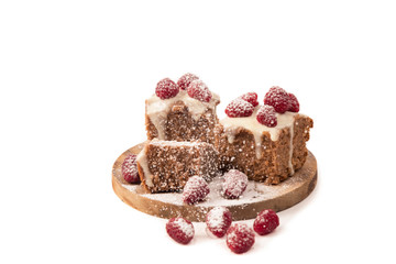 The chocolate cake decorated by white glaze and fresh berries of raspberry is strewed with icing sugar lies on a wooden round support. Separately on a white background. Horizontal shot. The place for 