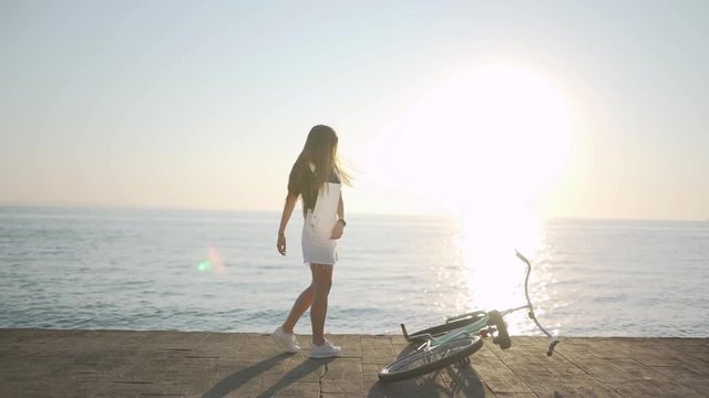 Young attractive woman with long hair is having a good time on sea at sunset or sunrise. Female with a bicycle