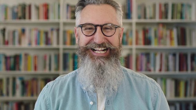 portrait of mature caucasian professor with beard laughing cheerful at camera in library bookshelf background wearing glasses