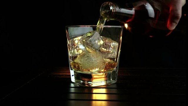 barman pouring whiskey in the glass with ice cubes on wood table and black dark background, focus on ice cubes, whisky relax time on warm atmosphere