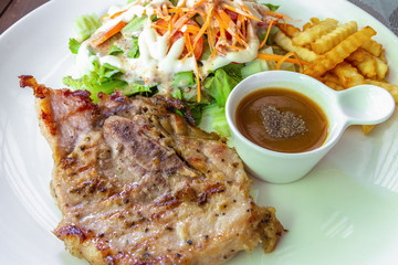 Medium grilled homemade pork steak cooked by black pepper and salt serve with gravy sauce, fresh salad and french fries on white dish.