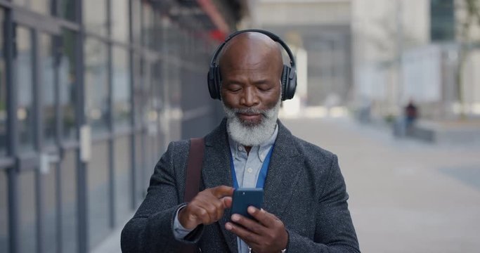 portrait senior african american businessman using smartphone in city enjoying listening to music wearing earphones browsing online messages texting on mobile phone slow motion