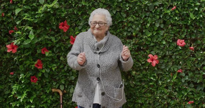 portrait of cheerful elderly woman dancing wearing earphones listening to music enjoying playful fun in beautiful green garden flower wall outdoors holding walking stick