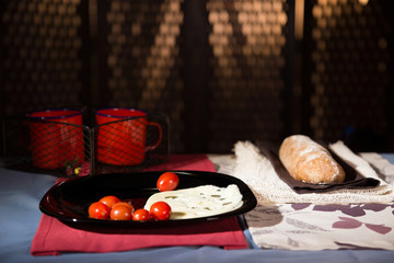 Cherry tomatoes and cheese on a black plate. Decorated background with a ciabatta bread and two red cups. 