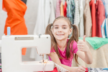 Happy little girl at workplace seamstress showing thumbs up. Hobby sewing concept