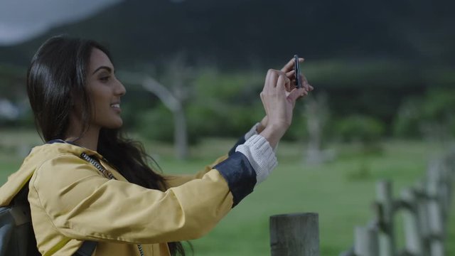 attractive young indian woman taking photo using smartphone camera technology enjoying sharing adventure travel hiker photographing green countryside outdoors on mobile phone