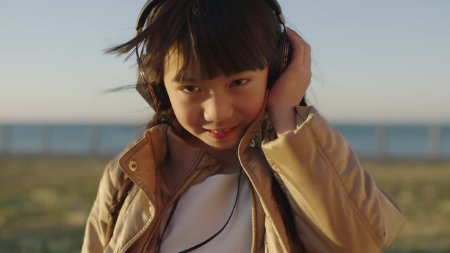 close up portrait of cute little asian girl wearing headphones smiling enjoying listening to music on seaside beach park at sunset