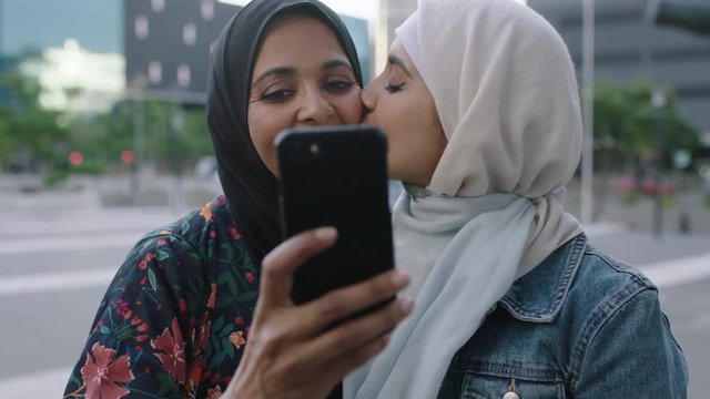 portrait of young muslim women posing daughter kisses mother on cheek taking selfie photo using smartphone camera technology in urban city background