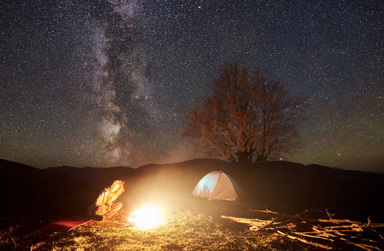 Camping night in mountains. Young tourist girl sits watching burning bonfire under deep dark starry sky with brightly lit tent and silhouette of big tree in background. Tourism and travel concept.