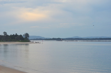 Late afternoon over a river inlet where it joins the ocean. A man is fishing from the beach. The sky is overcast.