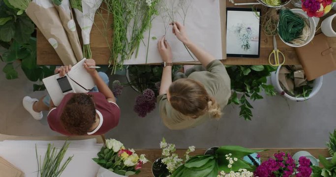 young florist business woman in startup flower shop making beautiful bouquet using tablet brainstorming creative ideas working together with colleague in successful store top view