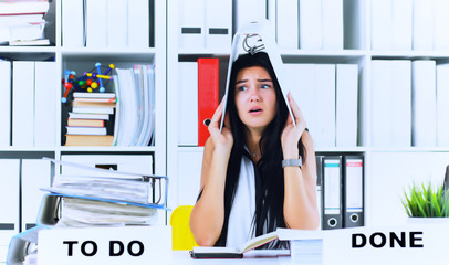 Funny overworked girl with a folder on her head sitting in the workplace cluttered with folders....