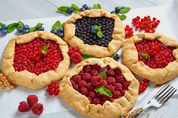 Various berries tarts with vintage knife and fork on the gray concrete background. Vegetarian healthy galette decorated with fresh blueberries, raspberries, red currants, mint leaves