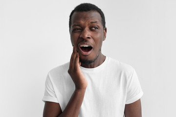Young african american man in blank white t shirt, isolated on gray background with mouth open, touching his face with expression of horrible suffer from health problem and aching tooth