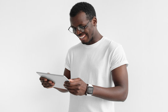 Indoor closeup of young handsome African man isolated on gray background dressed in t shirt, wearing round eyeglasses, looking at tablet screen with smile laughing while or surfing ne