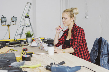 Beautiful young woman in checkered shirt sitting at wooden workbench with carpenter tools and working on laptop smiling