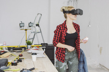 Cute blond young woman in casual clothes standing in virtual reality goggles headset at wooden workbench with tools and holding small white controller in hand © Dash