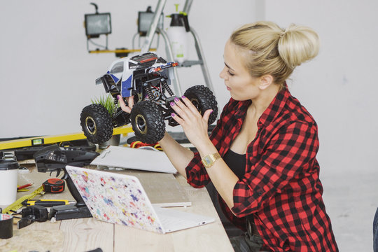 Side view of sitting at wooden workbench young blond female in checkered shirt concentrating on examining configuration of blue radio-controlled car in hands