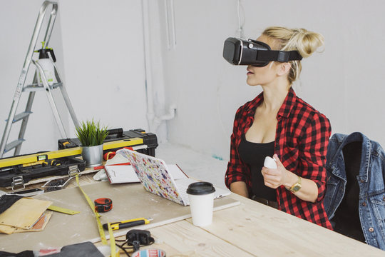 Young blond woman in casual clothes sitting at wooden workbench with tools and laptop and enjoying virtual reality experience in goggles headset holding joystick in hand.