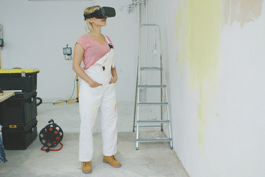 Smiling young woman in pink shirt and white overalls standing relaxed at workplace at half-painted yellow wall with hands in pockets in virtual reality goggles headset.