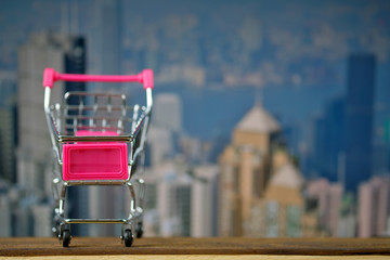 Shopping cart or supermarket trolley on top wooden working table with city city and office building background, business and financial concept.