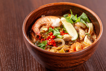 Asian Oriental Cuisine. Japanese spicy soup with seafood, shrimps, vegetables, lime, parsley, and hot pepper, in a plate of red clay, stands on a table in a restaurant. Copy space, selective focus