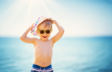 little boy smiling at the beach in hat with sunglasses