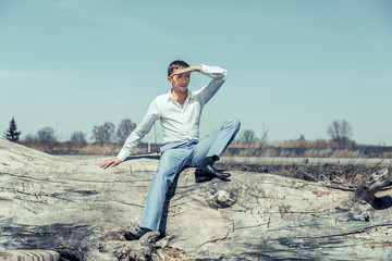 Fototapeta na wymiar A young guy in a white shirt and blue pants sits on a log against the blue sky and peers into the distance, covering his eyes with the hand from the sun.