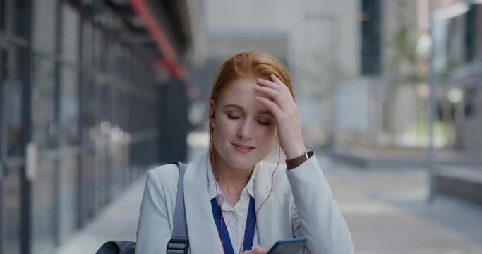 portrait happy young red head business woman using smartphone texting sending messages enjoying listening to music wearing earphones in relaxed urban city slow motion