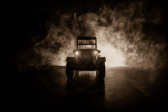 Silhouette of old vintage car in dark foggy toned background with glowing lights in low light.
