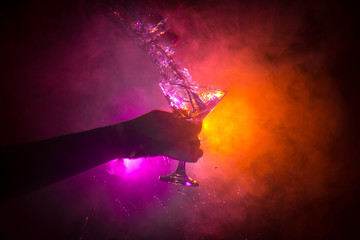 Martini cocktail glass in hand splashing on dark toned smoky background or colorful cocktail in glass with splashes and olives. Party club entertainment. Mixed light.