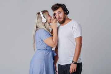 Naklejka premium Young cheerful and cheerful couple with wireless headphones, smiling against a gray background. The girl whispers the guy a secret in the ear. Club DJs